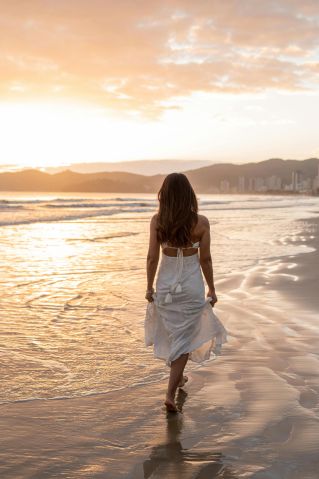 A woman in a white dress walks along a scenic beach during a golden sunset.